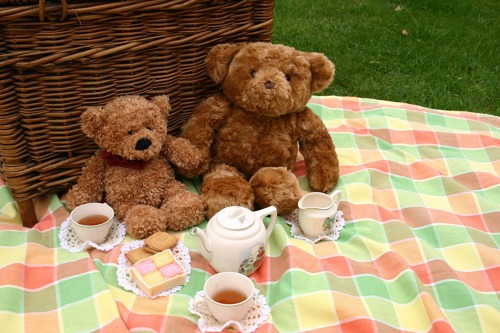 Teddy bears sat on picnic mat with a picnic basket behind them with tea and cake